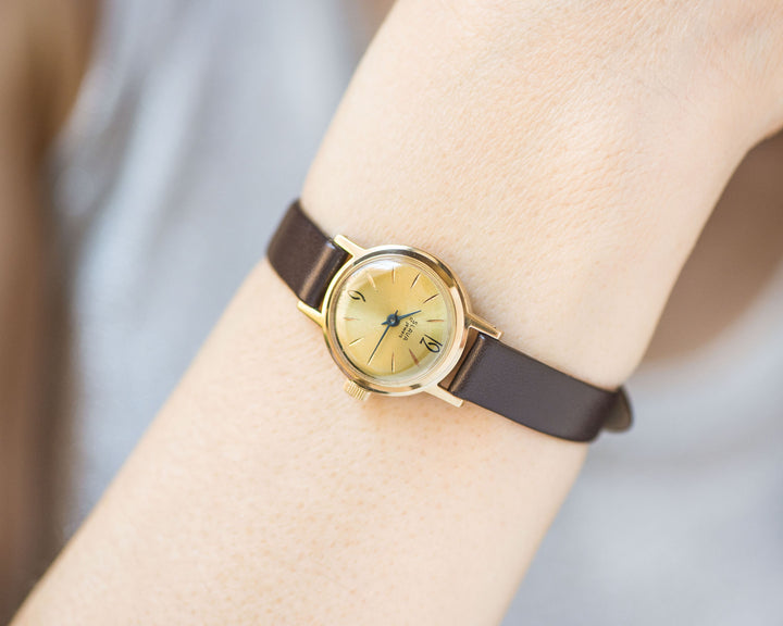 The tiny gold-plated watch on the woman’s wrist. The dial of the watch is with Arabic numerals, minimalist style, classic watch tiny little for every occasion. A piece of pre-owned vintage jewelry. You can make a better choice for the Planet.