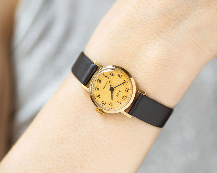 The tiny gold-plated watch on the woman’s wrist. The dial of the watch is with Arabic numerals, minimalist style, classic watch tiny little for every occasion. A piece of pre-owned vintage jewelry. The strap is dark brown premium leather.