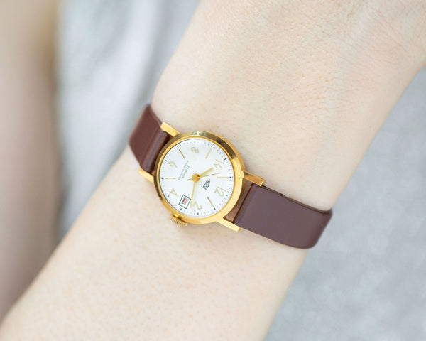 The tiny gold-plated watch on the woman’s wrist. The dial of the watch is with Arabic numerals, date window, minimalist style, classic watch tiny little for every occasion. Never been used vintage jewelry.