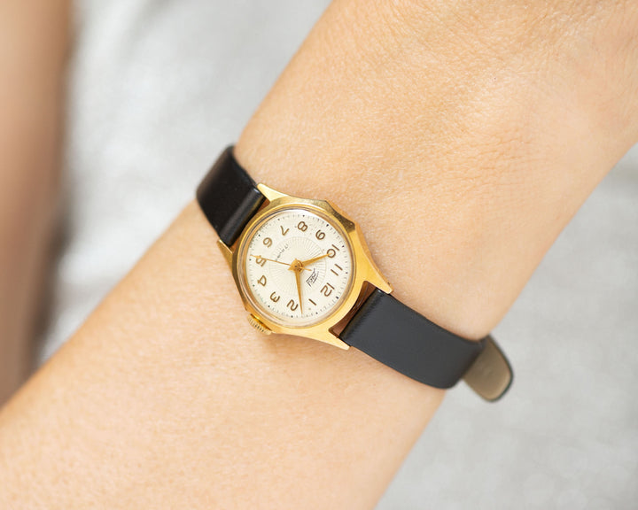 The tiny gold-plated watch on the woman’s wrist. The sunburts dial of the watch is with Arabic numerals, minimalist style, classic watch tiny little for every occasion. A piece of pre-owned vintage jewelry.